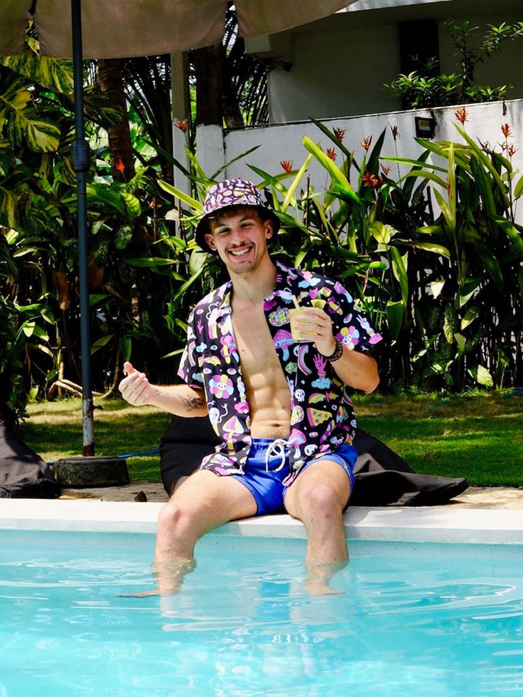 A smiling man wearing a colorful shirt, matching bucket hat, and blue swim trunks sits on the edge of a pool with his feet in the water, holding a drink, surrounded by lush greenery.